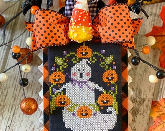 Peut inclure: Un motif de point de croix noir et blanc représentant un fantôme avec des citrouilles suspendues à des vignes. Le motif est encadré d'une bordure à carreaux noirs et blancs et d'un bord festonné noir. Un nœud à pois noirs et orange avec un dessus de maïs sucré est attaché au sommet.