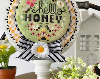 May include: A round, cross-stitched piece with the words "Hello Honey" surrounded by bees and flowers. It is mounted on a white stand and decorated with a black and white striped bow and a daisy. A rolling pin with the word "BEES" is in the foreground.
