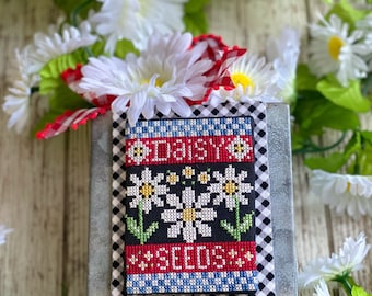 May include: A metal tin with a cross-stitch design featuring a black background with white daisies and the words "Daisy Seeds" in red. The tin is surrounded by white daisies.