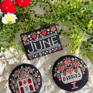 May include: A collection of cross-stitched items featuring a black background with strawberry and floral designs. One piece reads "JUNE", while two others depict a house and a basket of "BERRIES". Decorative crochet strawberries and greenery are also present.