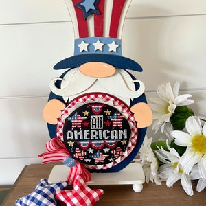 May include: Patriotic gnome decoration with a red, white, and blue hat, a white beard, and a blue and white oval with the text "I'm American" and stars. The gnome is holding a red, white, and blue gingham bow and is standing on a white base with two white legs. There are two red and blue gingham stars in the foreground.