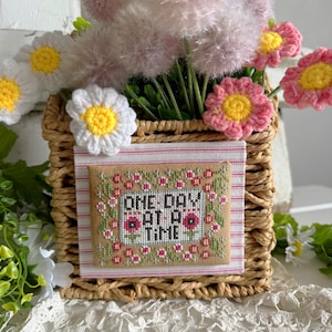 May include: A woven basket filled with pink and white flowers and greenery. A framed cross-stitch piece with the words "ONE DAY AT A TIME" is inside the basket. The basket is sitting on a white lace surface.