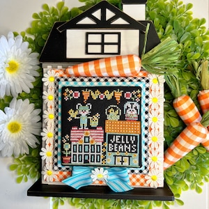 May include: A decorative Easter-themed house with a black roof, white walls, and a cross-stitch design. The cross-stitch features cartoon animals and the words "JELLY BEANS". The house is surrounded by a green wreath, daisies, and carrot decorations.