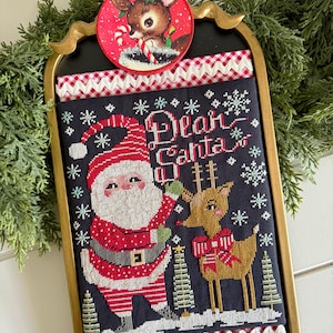 Peut inclure: Une broderie au point de croix de Noël encadrée représentant le Père Noël et un renne, avec les mots "Dear Santa". L'œuvre est encadrée en or et ornée d'une bordure vichy rouge et blanche. Un ornement rond avec un renne est attaché en haut.