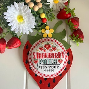 Può includere: Un ritaglio di fragola in legno rosso con un cerchio a quadri bianchi e rossi che dice "Strawberry Patch Pick Your Own" a punto croce. Il ritaglio è decorato con foglie verdi e un fiore giallo.