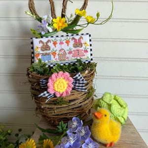 May include: A brown wicker basket with a pink and yellow flower and a black and white checkered ribbon. The basket is decorated with green moss and has two brown twig bunny ears. A cross-stitch design of bunnies and carrots is displayed in the basket. A yellow duck and purple flowers are in the foreground.