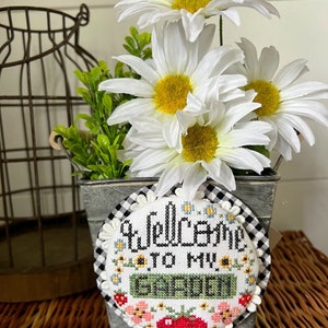 Pode incluir: Um balde de metal branco com um padrão xadrez preto e branco contém um buquê de margaridas brancas e um bordado de ponto de cruz redondo com o texto "Bem-vindo ao meu jardim".