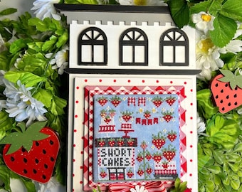 May include: A miniature wooden storefront with a red and white gingham roof and a sign that reads "Short Cakes". The storefront is decorated with strawberries and flowers.