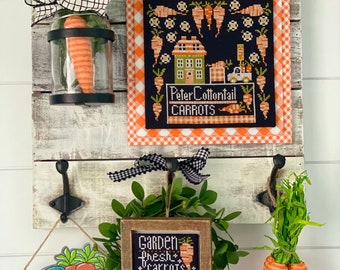 May include: A white wooden wall with three hooks. On the hooks are a framed cross-stitch with the text "Garden Fresh Carrots", a blue truck with the text "Hello Spring", and a bunch of orange carrots. There is also a framed cross-stitch with the text "Peter Cottontail Carrots" and a glass jar with a carrot inside.