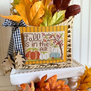 May include: A woven basket filled with colorful autumn leaves. The basket features a cross-stitched panel with the text "Fall is in the Air" and images of pumpkins and a tree. A black and white checkered ribbon adorns the basket.
