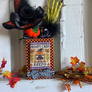 May include: A small wooden box with a black and white checkered pattern and a black bow. The box has a cross-stitch design of a witch holding a broom and the words "Broom Corn Seeds". The box is decorated with a witch's hat, a green hand, and a small orange pumpkin.