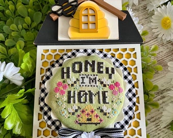 May include: A white wooden sign with a black and white checkered ribbon and a white daisy. The sign has a yellow honeycomb design and a green cross-stitch circle with the text "Honey I'm Home" and a beehive with bees.