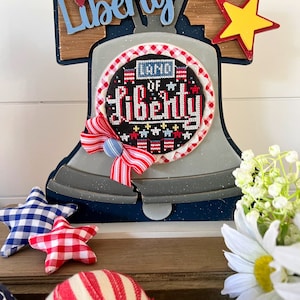 May include: A wooden bell with a blue and white star-shaped design. The bell is decorated with a red, white, and blue cross-stitch design that reads "Land of Liberty". The bell is surrounded by red, white, and blue stars and a white daisy.