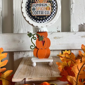 May include: A white wooden stand with a stack of three orange pumpkins. The top pumpkin has a green stem and a heart cut out. The stand has a round white base with a scalloped edge. A round white frame with a black and white checkered background and a cross-stitch design that says "Pumpkin Spice Coffee Co." is attached to the top of the stand.