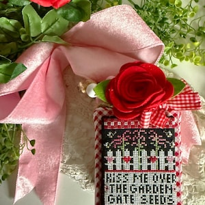 May include: A handmade cross-stitch piece with the text "Kiss Me Over The Garden Gate Seeds." The piece is framed with a red and white checkered pattern. A red felt rose and pink ribbon are attached to the top, with greenery and a red rose in the background.