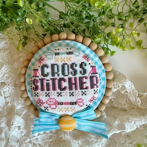 May include: A round, cross-stitched design with the words "CROSS STITCHED" in black and pink. The design is surrounded by a blue and white gingham pattern, wooden beads, and a blue ribbon with a yellow button.