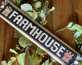 Pode incluir: Uma placa preta e branca com a palavra "FARMHOUSE" em letras brancas. A placa é enquadrada em madeira e tem uma borda xadrez preta e branca. Há duas pequenas imagens bordadas na placa, uma de uma casa e outra de um celeiro.