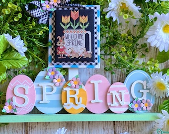 Op de afbeelding: Een decoratief bord met een lentethema met een kruissteekontwerp met tulpen en de woorden "WELCOME SPRING". Het bord bevat een ruitlint en een houten "SPRING" bord met pastelkleurige paasei-accenten en bloemversieringen.