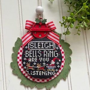 May include: A round, black and white Christmas ornament with the text "Sleigh Bells Ring Are You Listening" and a red and white gingham border. The ornament is decorated with a red bell and a red and white striped bow.