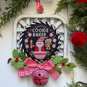 Puede incluir: Un marco de madera blanco con un fondo a cuadros blanco y negro. Una cinta de cuadros rojos y blancos está atada alrededor del marco. Una campana roja con una estrella cuelga de la cinta. Un diseño de punto de cruz blanco y negro con el texto "Cookie Baker" está dentro del marco. El diseño presenta una figura de Santa Claus y un pequeño hombre de jengibre.