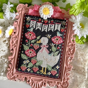 May include: A framed cross-stitch featuring a white duck with a pink bow, surrounded by red flowers and strawberries. The word "SWEET" is stitched across the top. The frame is pink with a gingham ribbon and daisy accent.