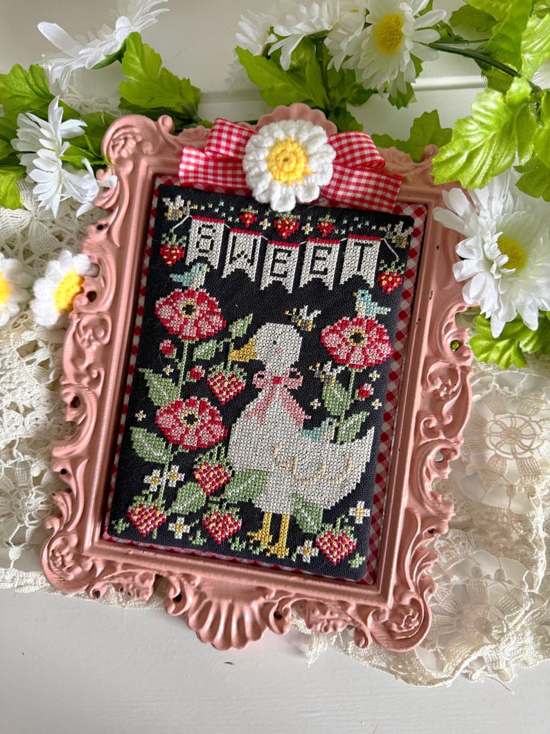 May include: A framed cross-stitch featuring a white duck with a pink bow, surrounded by red flowers and strawberries. The word "SWEET" is stitched across the top. The frame is pink with a gingham ribbon and daisy accent.