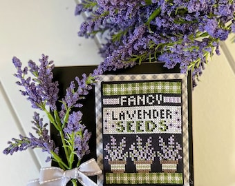 May include: A black framed cross-stitch with a white and green checked border. The cross-stitch features the text "Fancy Lavender Seeds" and three potted lavender plants. The frame is sitting on a white surface with a sprig of lavender flowers and a grey ribbon.