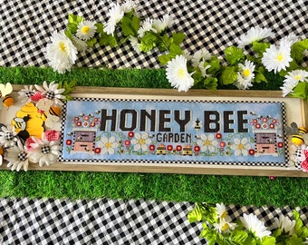 May include: A framed cross-stitch design with the text "Honey Bee Garden" surrounded by bees, flowers, and beehives. The frame is on a green grass surface with white and black checkered fabric in the background.