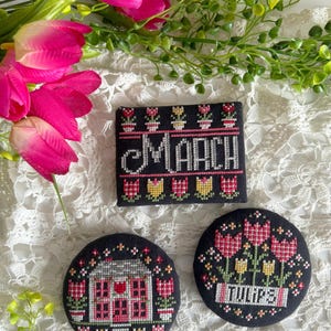May include: Black cross-stitch pieces with floral and house designs. One piece reads "MARCH" in white, with tulips above and below. Two round pieces feature a house and the word "TULIPS". Pink tulips and greenery are in the background.