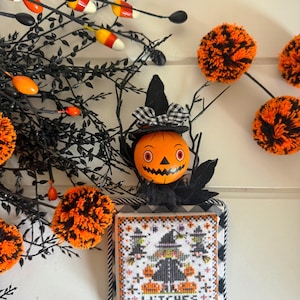 Könnte beinhalten: Ein quadratisches Kreuzstich-Ornament mit einem Hexen- und Kürbismotiv und dem Text WITCHES, gekrönt von einem schwarzen Hexenhut, einer schwarz-weiß karierten Schleife und einem runden orangefarbenen Kürbisgesicht mit schwarzen Details.