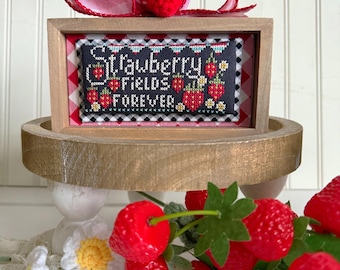 May include: A framed cross-stitch design with a black background and red and white strawberries. The text reads "Strawberry Fields Forever". The frame is sitting on a wooden tiered stand with a red and white gingham bow and a crocheted strawberry on top.