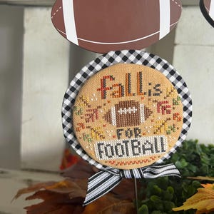 May include: A decorative fall-themed sign with a football and the text "Fall is for Football." The sign features a brown football shape and a round stitched design with a black and white checkered border. A black and white striped bow is attached.