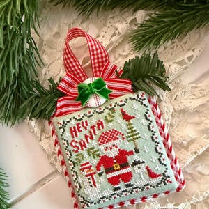 May include: A square cross-stitch ornament featuring a light green background with a Santa Claus figure, a small tree, and the text "HEY SANTA". The ornament is accented with a red and white gingham ribbon, a small green bow, and a white button.