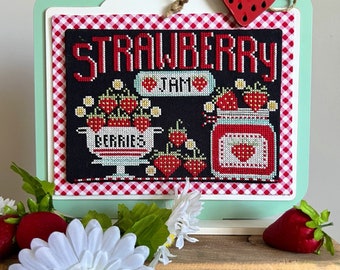 May include: A cross-stitch embroidery design featuring a jar of strawberry jam with the text "Strawberry Jam" and "Berries" in red and white lettering on a black background. The design is framed with a red and white gingham border and displayed in a white and green mason jar frame.
