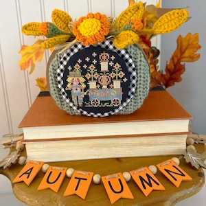 May include: A sage green crocheted pumpkin featuring a black and white gingham border and a cross-stitch design of a scarecrow and wagon. It is topped with crocheted orange flowers and leaves, resting on two books with an orange wooden banner reading AUTUMN.