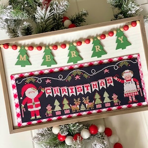 May include: A framed cross-stitch design featuring a Christmas scene with Santa Claus, elves, reindeer, and a banner that reads "CHRISTMAS". The frame is decorated with a garland of green pine branches and red and white beads.