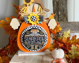 May include: A wooden scarecrow figurine with a pumpkin-shaped body and a black and white checkered pattern. The scarecrow is wearing a brown hat and a yellow bow. The pumpkin has a stitched circle with the text "Scarecrow Festival".