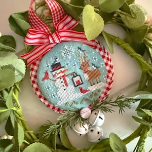 May include: A round, cross-stitched Christmas ornament featuring a snowman and reindeer in a snowy scene. The ornament has a light blue background, red and white gingham trim, and a red and white striped ribbon. White bells and greenery complete the festive design.
