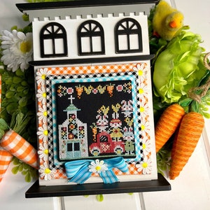 Könnte beinhalten: Ein dekoratives Kreuzstichbild zum Thema Ostern, gerahmt in einem weißen, hausförmigen Holzrahmen mit schwarzen Akzenten. Der Kreuzstich zeigt eine Kirche und Hasen in einem roten Lastwagen. Eine blaue Schleife mit einer Gänseblümchen ist am Rahmen befestigt.