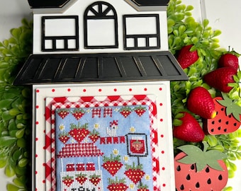 May include: A white wooden house with a black roof and windows, decorated with a red and white gingham fabric and a cross-stitch design of a strawberry farm with the text "See Jam". The house is surrounded by green leaves and red strawberries.