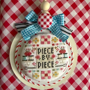 May include: A handmade cross-stitch ornament with the text "PIECE BY PIECE". The ornament features a white background with colorful floral and sewing-themed designs. It is adorned with a red and white gingham bow and a wooden bead.