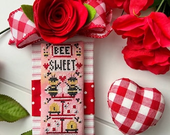 May include: A cross-stitch ornament with a pink background and the text "Bee Sweet" in black. Two bees are shown on a branch with a heart between them. The ornament is surrounded by red and white gingham ribbon and red roses.