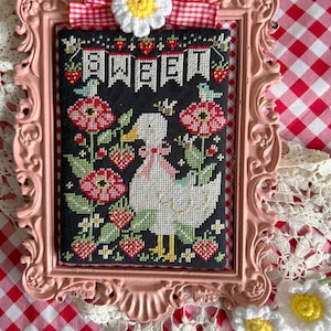 Könnte beinhalten: Ein gerahmtes Kreuzstichbild mit einer weißen Ente mit Krone, umgeben von roten Blumen und Erdbeeren. Der schwarze Hintergrund trägt das Wort "SWEET" in weißen Buchstaben. Der rosa Rahmen ist mit einer rot-weiß karierten Schleife und einer gehäkelten Gänseblümchen verziert.