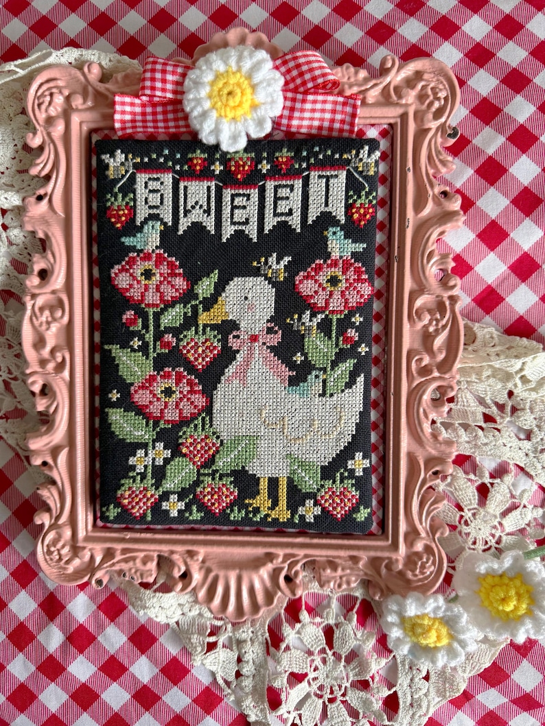 May include: A framed cross-stitch featuring a white duck wearing a crown, surrounded by red flowers and strawberries. The black background has the word "SWEET" in white letters. The pink frame is adorned with a red and white checkered bow and a crocheted daisy.