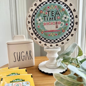 May include: A white wooden stand with a scalloped edge and a black and white checkered background. The stand has a cross-stitch design of a teacup with the words "Tea Time" in a floral wreath. The stand is on a wooden table with a sugar container and tea bags.