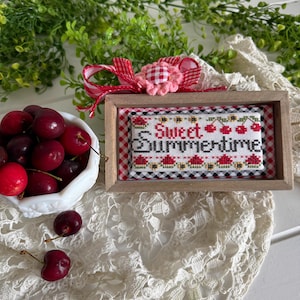 May include: A framed cross-stitch piece with the words "Sweet Summertime" and cherry designs. The frame is wooden, and the background is a red and white gingham pattern. A bowl of fresh cherries sits nearby, with a red and white gingham bow.