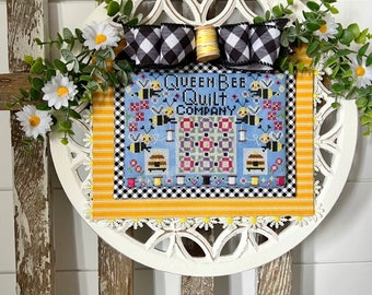 Pode incluir: Uma moldura circular de madeira branca com uma fita xadrez preta e branca e um carretel de fio amarelo. A moldura contém uma peça de ponto cruz com fundo azul e o texto "Queen Bee Quilt Company" e um desenho de abelhas e flores.