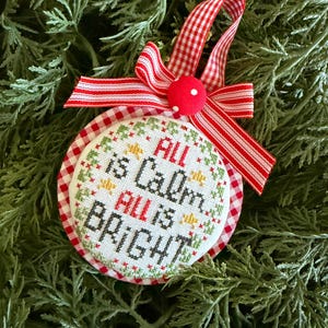 May include: A round Christmas ornament with the words "All is Calm, All is Bright" embroidered in red and green. The ornament has a red and white gingham border and is topped with a red bow and a red pom-pom. The ornament is displayed on a bed of green pine needles.