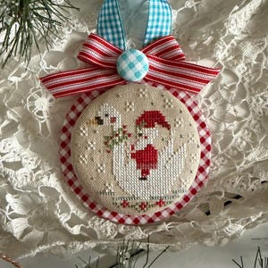 May include: A handmade Christmas ornament with a cross-stitch design. The ornament features a round shape with a red and white gingham border. The center shows a snowman and a figure in a red Santa suit. A blue and white gingham ribbon and a red and white striped bow complete the design.