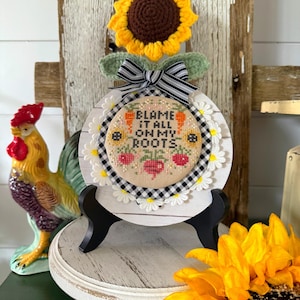 May include: A round, white wooden sign with the text "BLAME IT ALL ON MY ROOTS" in a cross-stitch design. The sign is decorated with a black and white checkered pattern, daisies, and a crocheted sunflower. A ceramic rooster is in the background.